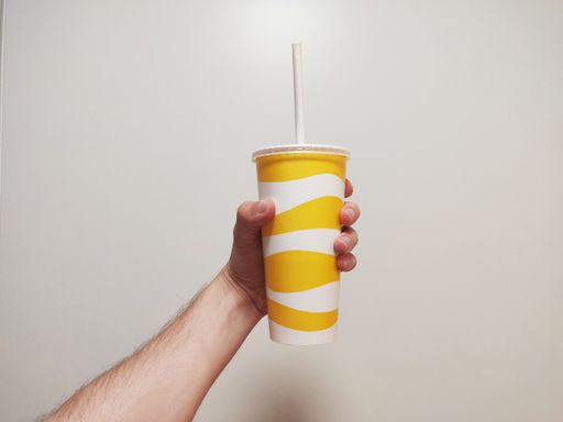 A closeup of a male hand holding a tall drink in a white and yellow plastic cup in front of a wall