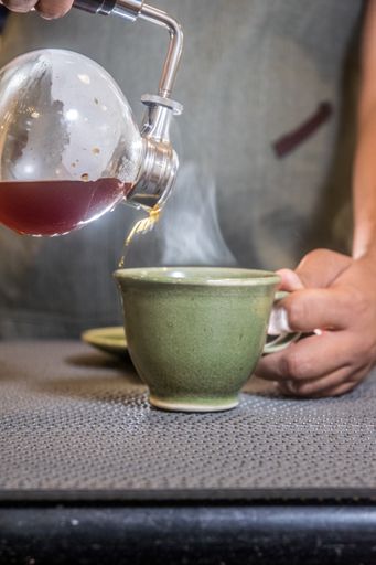 A vertical shot of a man pouring coffee in the cup from a siphon coffee maker