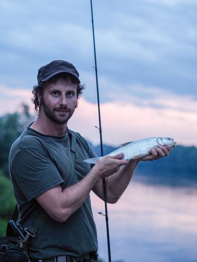 A vertical shot of a Caucasian fisherman showing his catch holding his rod standing on a bay