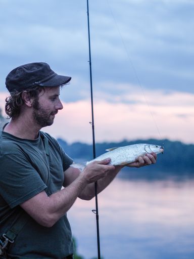 A vertical shot of a Caucasian fisherman showing his catch holding his rod standing on a bay