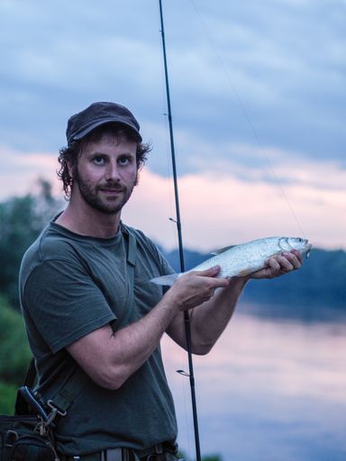 A vertical shot of a Caucasian fisherman showing his catch holding his rod standing on a bay