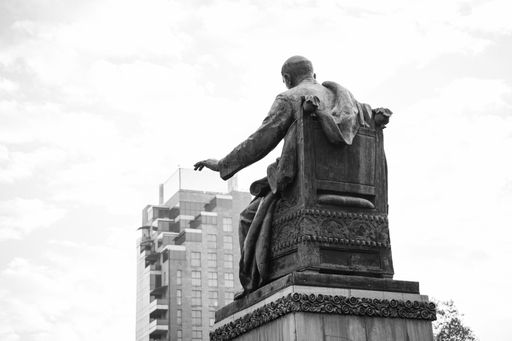 Black and white photo of a statue on a throne in front of a tall building with a cloudy sky