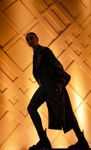 Silhouette of a stylish person in a long coat against a geometric, illuminated wall, showcasing modern fashion and architecture.