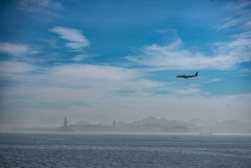 Airplane flying over ocean with distant cityscape and mountains under a clear blue sky.