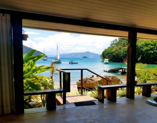 Tropical beachfront view from a shaded patio, featuring sailboats and lush greenery on a sunny day.