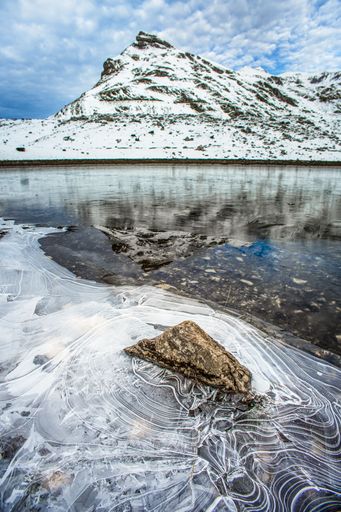 A snow-covered mountain with a lake in the foreground under a cloudy blue sky