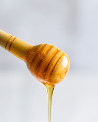 A vertical closeup of a wooden honey dipper with sweet honey dripping from it on a blurry background
