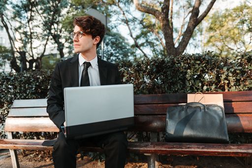 A serious young Caucasian guy dressed professionally sitting on a bench using his laptop