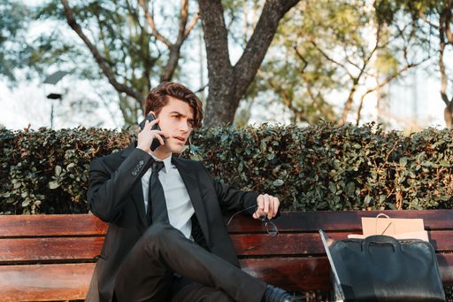 A serious young Caucasian guy dressed professionally sitting on a bench talking on his phone