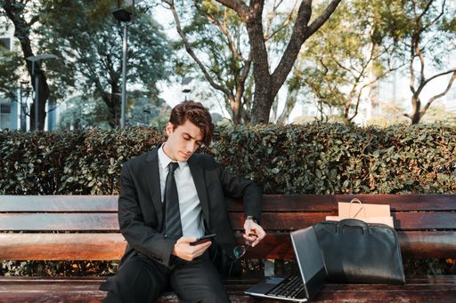 A serious young Caucasian guy dressed professionally sitting on a bench using his smartphone