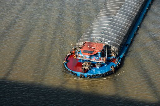 Aerial view of a cargo boat navigating a river with shadow patterns on the water surface.