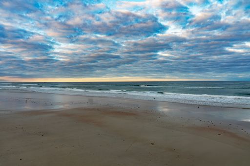 Serene beach scene with gentle waves under a dramatic cloudy sky at sunset, reflecting on wet sand.
