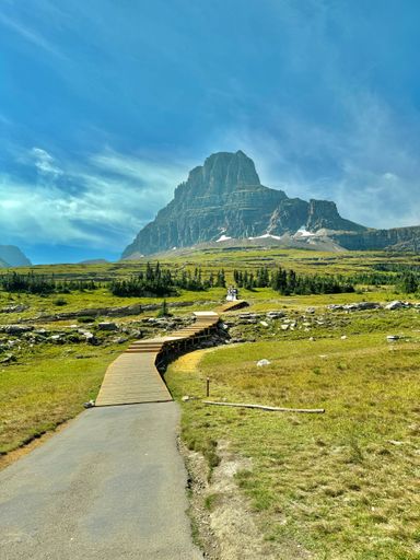 Scenic view of a mountain with a wooden path leading through grassy fields under a clear blue sky in Glacier National Park.