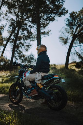 A person in a helmet sits on a motorcycle in a forest, with sunlight filtering through the trees, creating a serene outdoor scene.