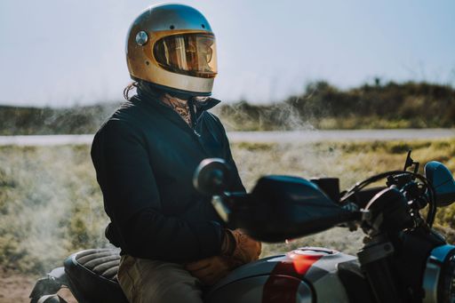 A motorcyclist in a golden helmet sits on a bike by a country road on a sunny day, exuding adventure and freedom.