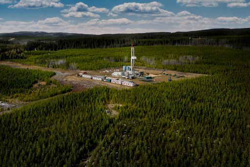 Aerial view of an oil drilling rig surrounded by dense forest under a cloudy sky, showcasing industrial activity in a natural setting.
