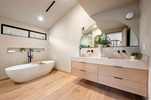 Modern bathroom interior with a freestanding tub, wooden vanity, and large mirror, featuring natural light and minimalist decor.