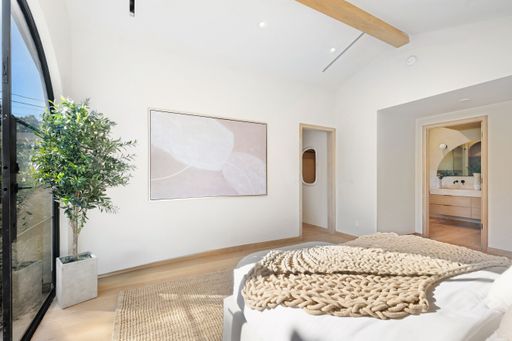 A bright, minimalist bedroom interior with natural light, featuring a large window, potted plant, and cozy decor.