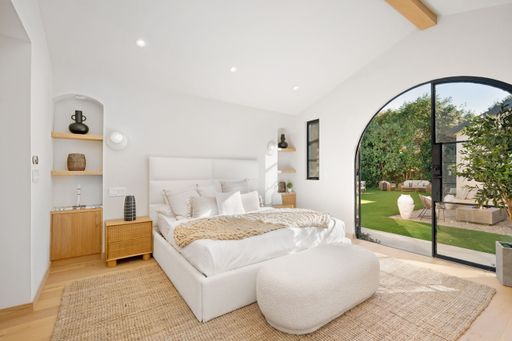 Modern bedroom with natural light, cozy decor, and garden view through large arched window, featuring neutral tones and stylish furnishings.