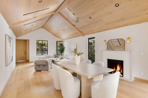 Modern living room with light wood floors, vaulted ceiling, and a cozy fireplace, featuring stylish furniture and large windows.