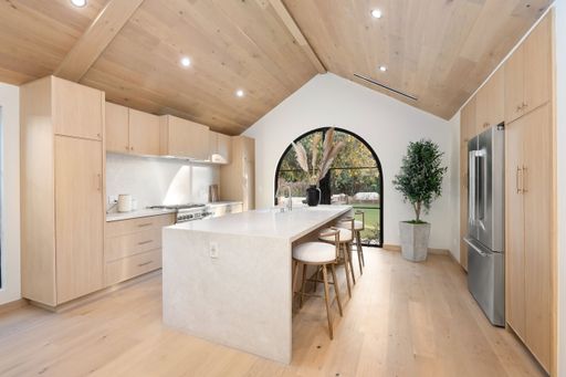 Modern kitchen interior with light wood cabinetry, large island, and arched window overlooking a garden.