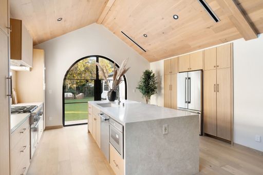 Modern kitchen interior with wooden cabinetry, high ceiling, and large arched window overlooking a garden.