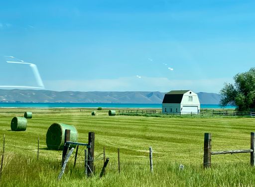 Scenic view of a rural landscape with hay bales, a white barn, and a distant lake under a clear blue sky.