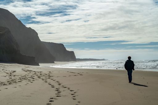 Person walking along a scenic beach with cliffs and ocean waves under a partly cloudy sky.