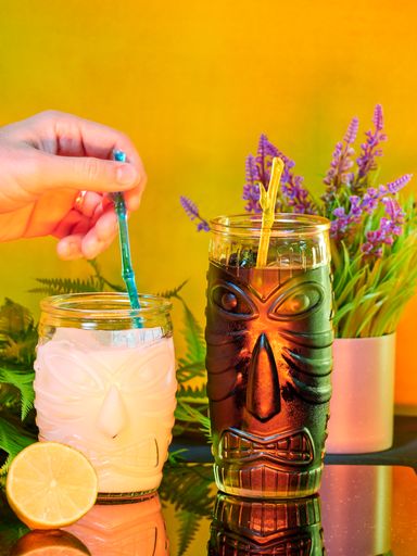 A vertical shot of a hand mixing cocktail with a straw in a fancy glass with a tiki face