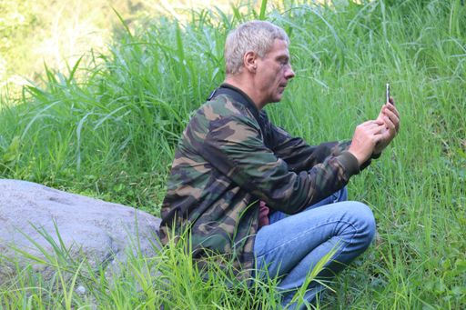 Profile view from a Caucasian mature adult man, using his smartphone, while sitting on a stone in a meadow. Low angle view.