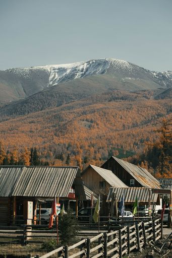 Scenic view of a rustic village with wooden houses, autumn trees, and snow-capped mountains under a clear sky.