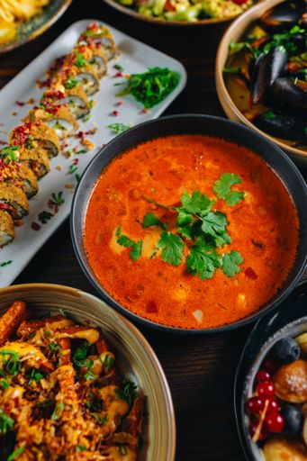 A vibrant array of Asian cuisine including spicy soup, sushi rolls, and seafood, garnished with fresh herbs on a wooden table.