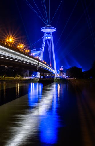 Night view of the UFO Bridge in Bratislava, Slovakia, with vibrant blue lights reflecting on the river.