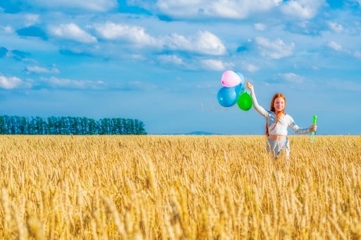 A young happy red-haired girl with multi-colored balloons in her hand runs along among a yellow wheat field against a blue cloudy summer sky.
