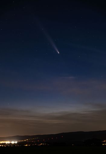 Comet streaks across a starry night sky above a distant cityscape, creating a serene and captivating celestial scene.