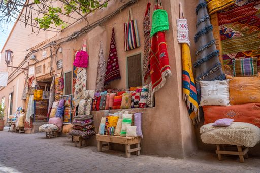 Colorful Moroccan textiles and pillows displayed on a narrow street in Marrakech, showcasing traditional patterns and vibrant colors.