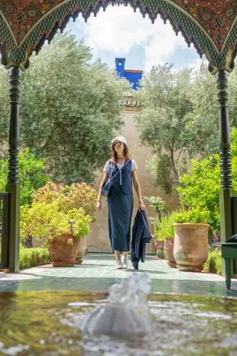 A woman in her 30s walks through a lush garden with a traditional archway, featuring vibrant greenery and a fountain in Marrakesh, Morocco.
