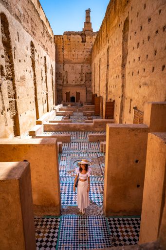 A woman in a white dress and hat explores the historic ruins of El Badi Palace in Marrakech, Morocco, under a clear blue sky.