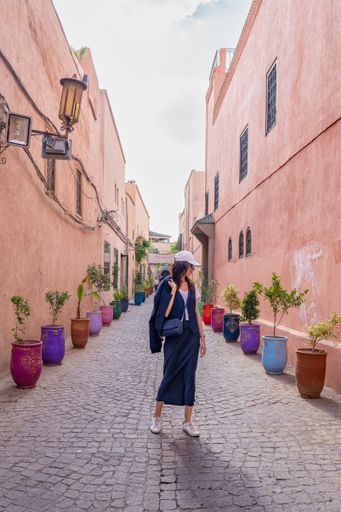 A young woman in casual attire walks down a colorful alley in Marrakech, Morocco, surrounded by vibrant potted plants and traditional architecture.