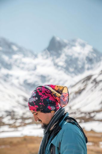 Man in colorful headscarf and jacket against snowy mountains, clear sky, enjoying outdoor adventure.