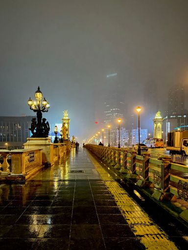 A misty evening view of a city bridge with ornate lamps and wet pavement, reflecting city lights, creating a moody urban atmosphere.