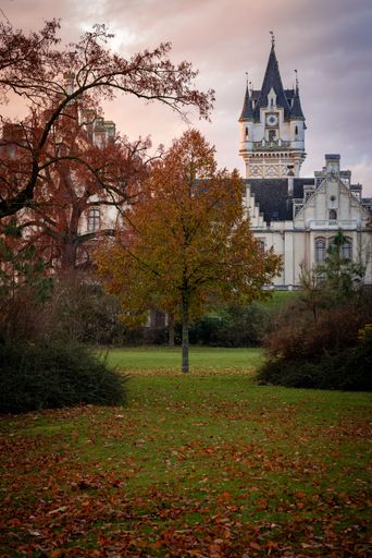 Charming autumn scene of a historic castle with a clock tower, surrounded by colorful trees and fallen leaves in a tranquil park setting.