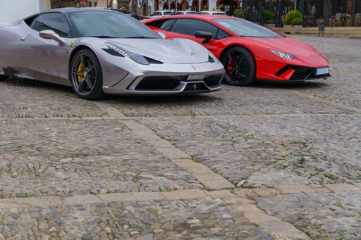 Two sleek sports cars, a silver Ferrari and a red Lamborghini, are parked side-by-side on a cobblestone street.