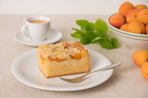A slice of apricot crumble cake on a plate with a fork, accompanied by a cup of coffee and fresh apricots on a table.