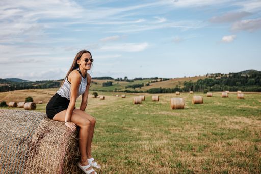A young woman in casual summer attire sits on a hay bale in a scenic rural field under a blue sky, enjoying a sunny day.