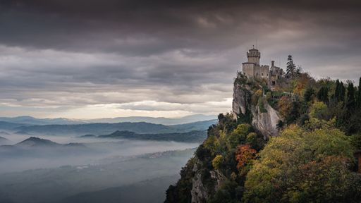 A dramatic view of a medieval castle perched on a cliff with a misty valley below and a stormy sky above in San Marino.