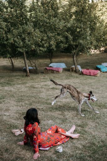 A vertical shot of a barefoot woman wearing red dress sitting on grass and watching her dog play in the field