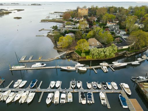 Aerial view of a marina with boats docked along the shoreline in a picturesque residential area, surrounded by lush greenery and calm waters.