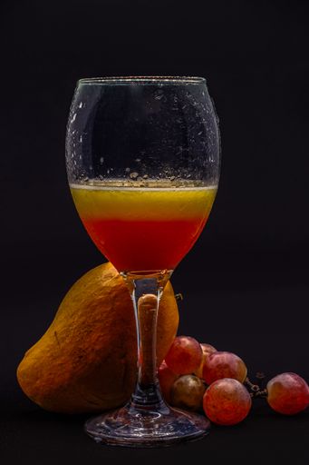 A vertical shot of a glass filled up with red and yellow liquid with mango and grapes in the background