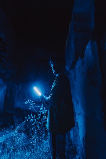 A young attractive Spanish man with oversized clothes holding a blue light in a ruined building